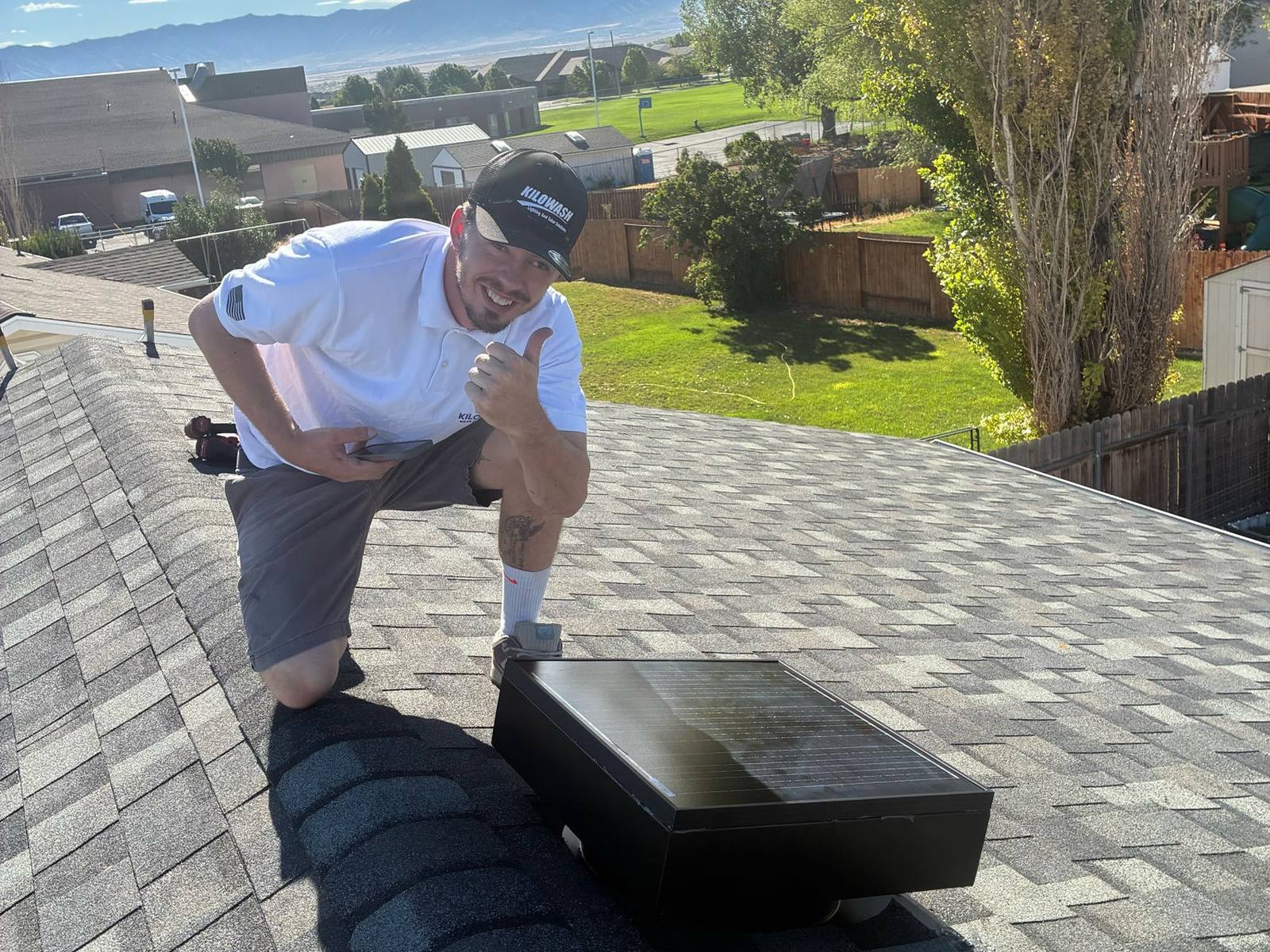Kilowash installer in a branded cap giving a thumbs up beside a solar attic fan he has just installed on a shingle roof.