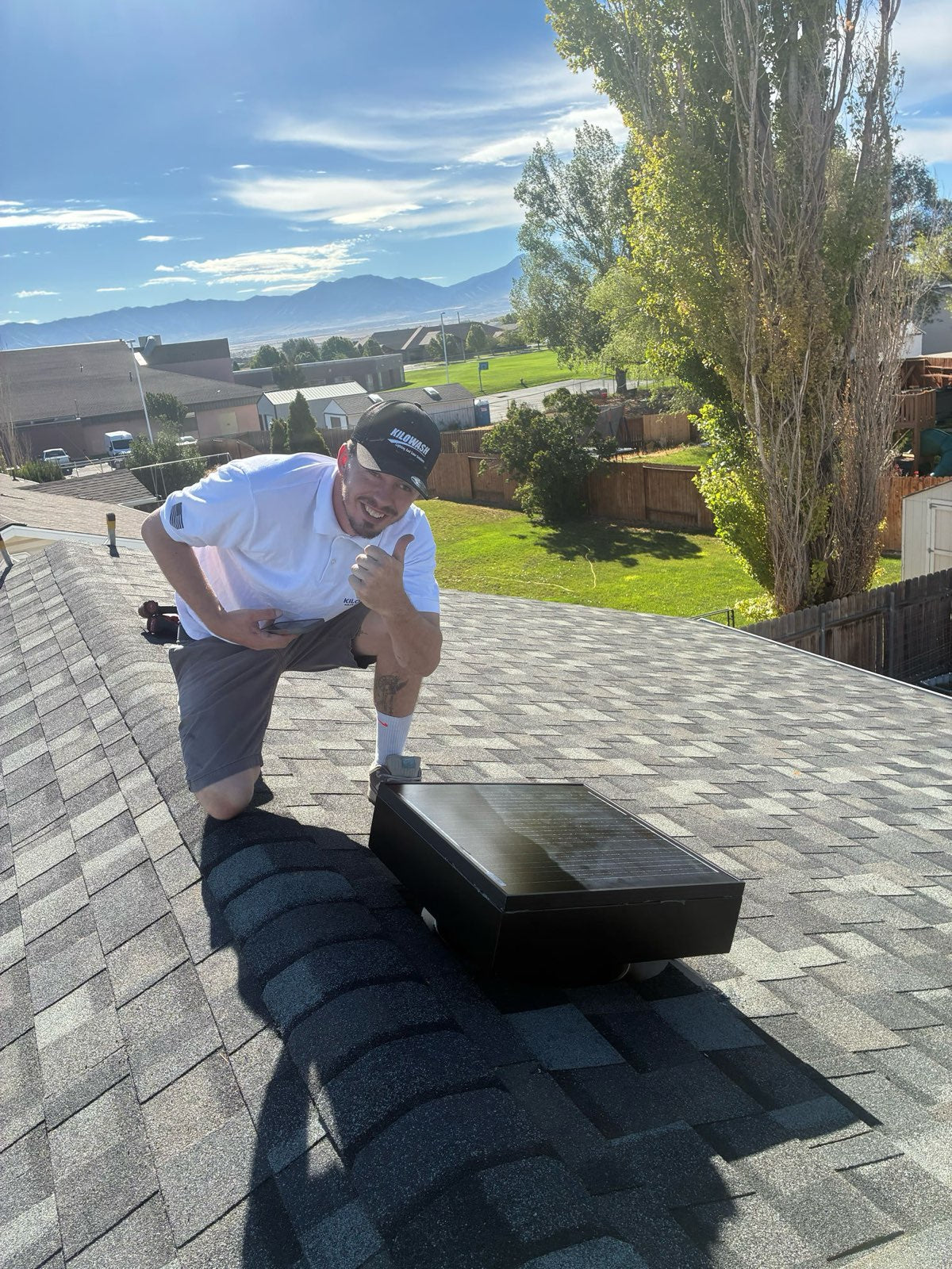 Kilowash installer in a branded cap giving a thumbs up next to a newly installed solar attic fan on a shingle roof, mountains behind.